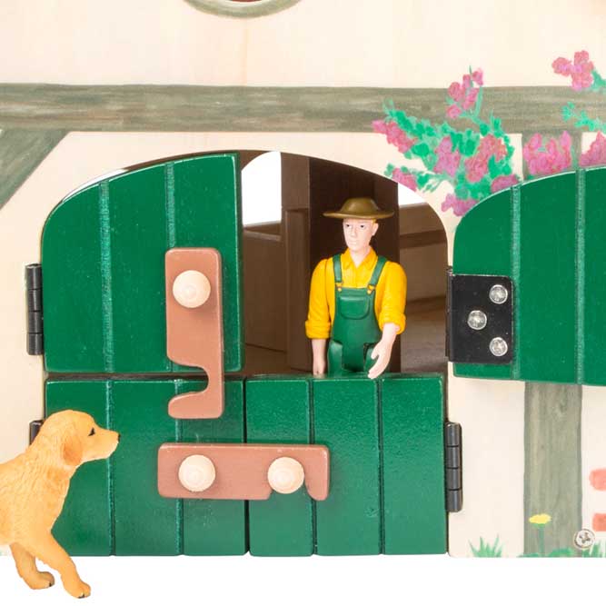La construcción de madera de la granja juguete está dispuesta en forma de ángulo  y equipada con un techo plegable. Detalle de la puerta del establo en color verde con el perro y el granjero.