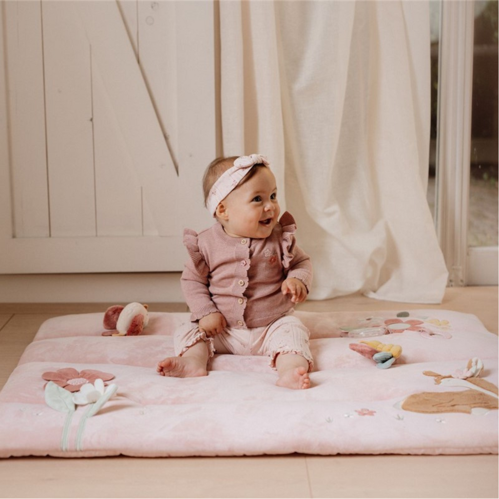 Bébé souriant assis sur un tapis de jeu rose avec des textures, des fleurs et des animaux