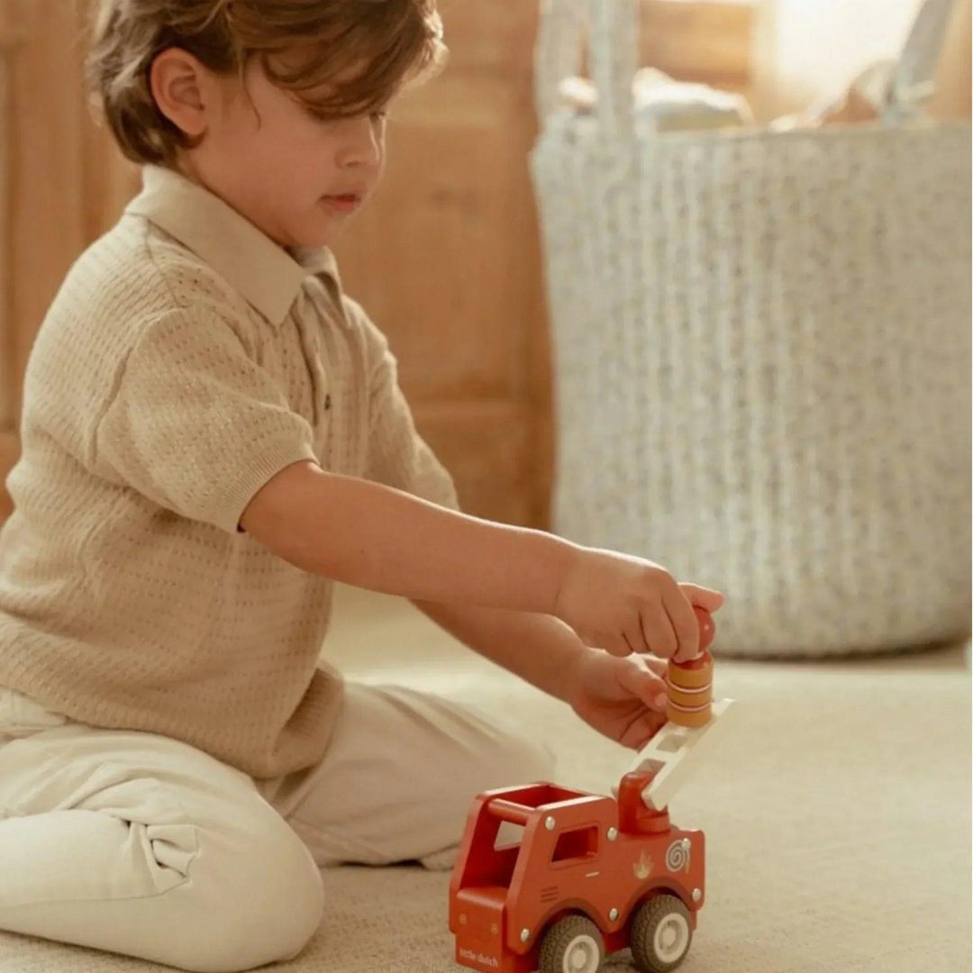 Petit garçon jouant avec un camion de pompiers en bois Little Dutch dans une chambre d'enfant éclairée.