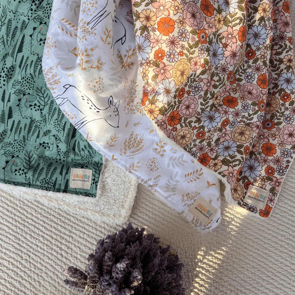 Three baby blankets with nature-inspired prints: a green one with animal drawings, a white one with a design of fawns and plants, and a floral one in warm tones.