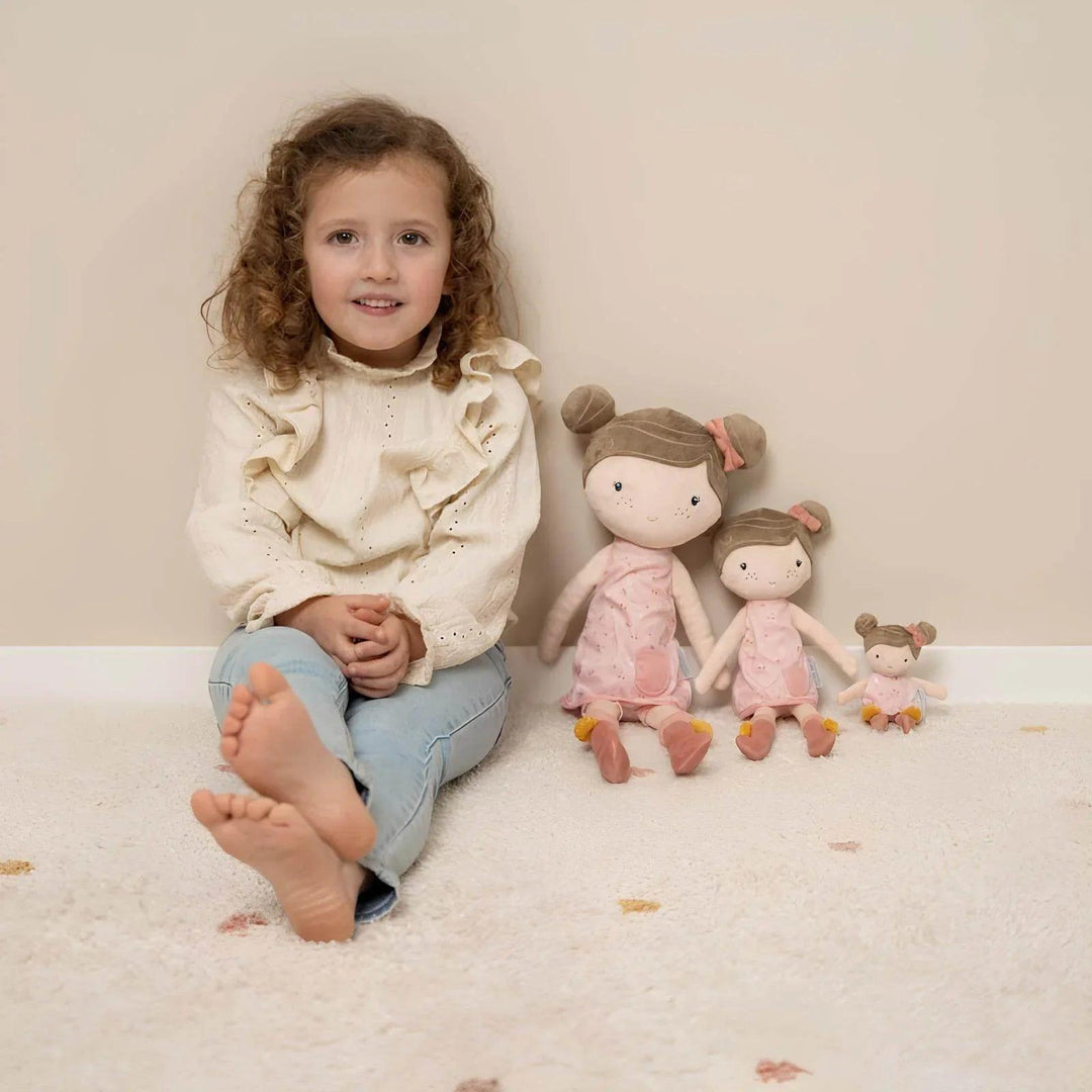 Little girl smiling next to Little Dutch dolls of different sizes wearing pink dresses and bows in their hair