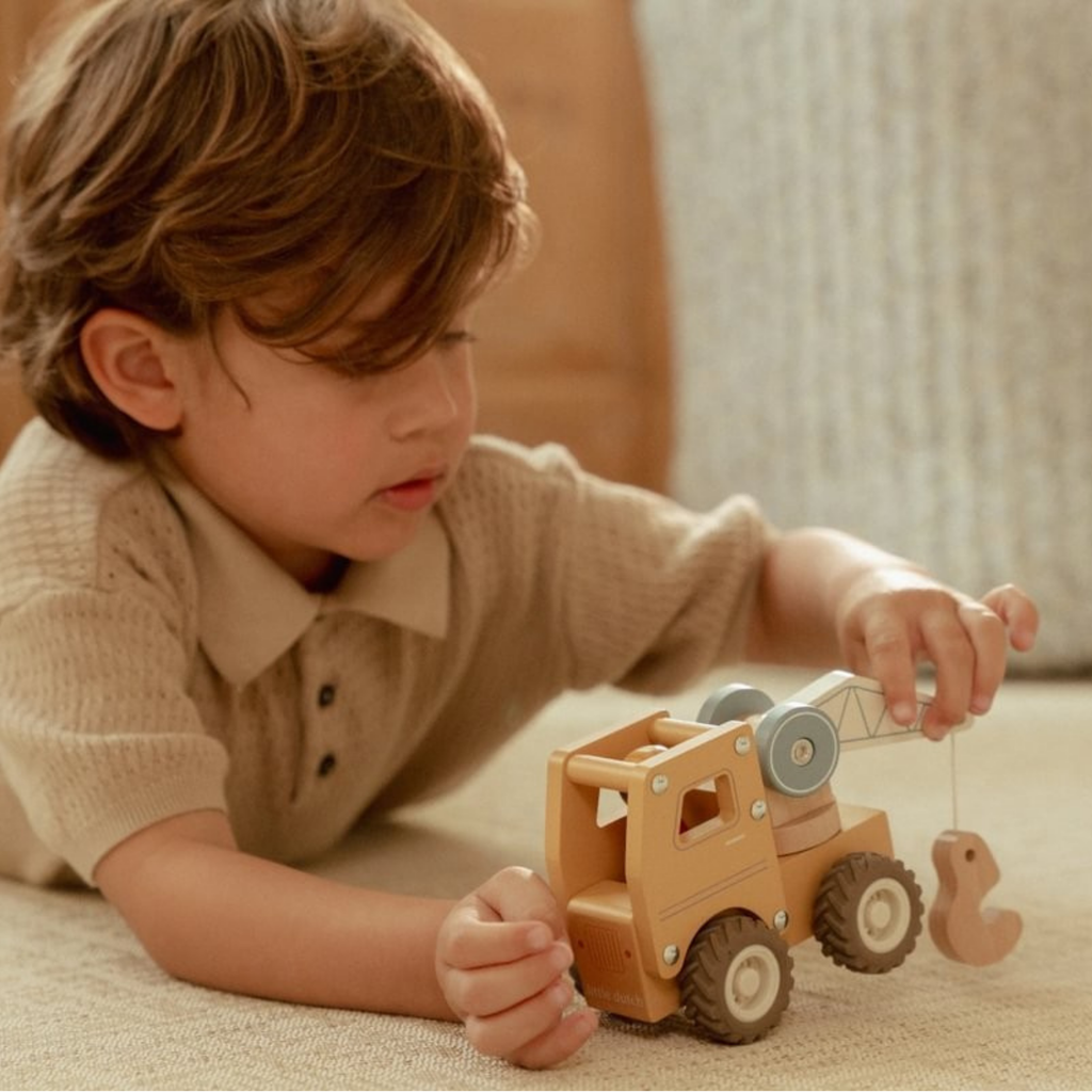 Niño jugando con una grúa de madera Little Dutch sobre una alfombra beige, moviendo el gancho del vehículo