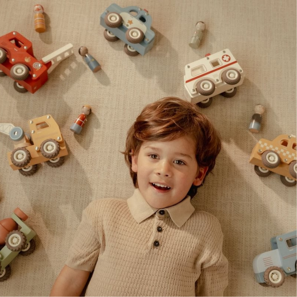 Niño sonriendo acostado en el suelo rodeado de vehículos de madera Little Dutch, incluyendo excavadora, ambulancia y camión de bomberos