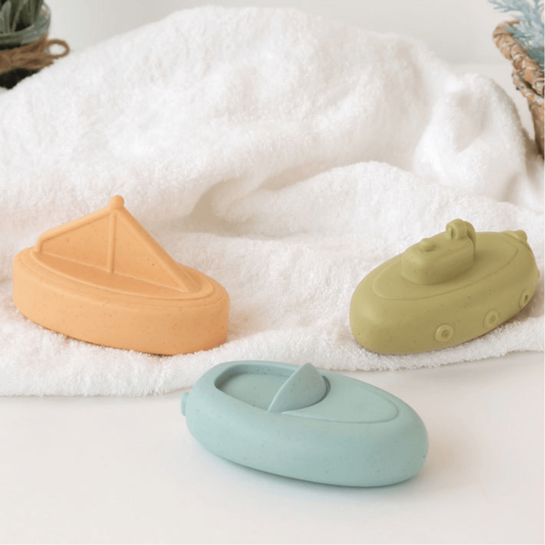 Tres juguetes de baño para bebé en forma de barco de silicona en colores pastel sobre fondo blanco.