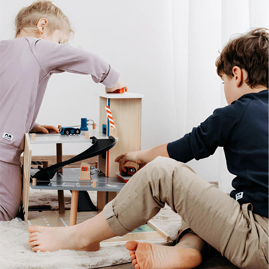 Due bambini giocano con un parcheggio in legno dotato di rampe, macchinine e accessori, che stimolano il gioco creativo.