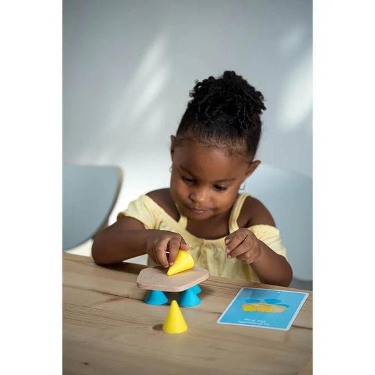 Fille jouant avec le jeu de construction Piks, empilant des pièces en bois et des cônes jaunes et bleus sur une table en bois