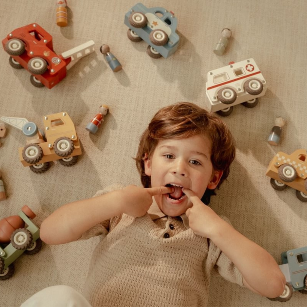 Niño acostado en el suelo rodeado de vehículos de madera Little Dutch, incluyendo grúa, ambulancia y camión de bomberos